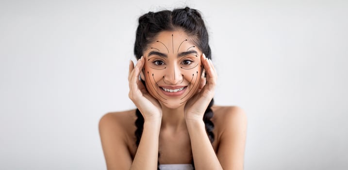 Indian woman is happily engaging in her skincare routine, applying products with care. She has a bright smile and is surrounded by an empty, well-lit space for her self-pampering session. photo