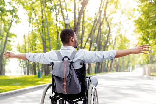 Back view of young disabled black man in wheelchair spreading his arms, feeling happy and free on walk at city park. Full length of handicapped guy with backpack going on trip outdoors photo