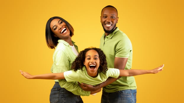 Family bonding concept. Joyful black man and woman holding daughter on hands, having fun over yellow studio wall. Smiling girl pretending she is flying, spreading hands, imitating plane photo