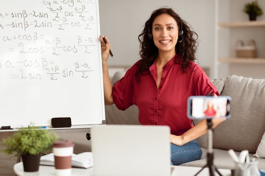 Online Learning Concept. Portrait of smiling lady teacher in headset sitting near whiteboard, explaining math formulas to students. Excited woman posing and looking at camera, recording class photo