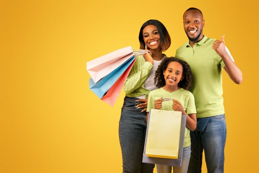 Black friday concept. Happy black parents and cute daughter holding shopper bags, man showing thumb up while posing together over yellow background. Family enjoying seasonal sales photo