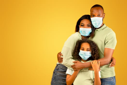 Young african american parents and their daughter wearing protective medical masks, embracing and looking at camera over yellow background. Black family following measures against coronavirus photo