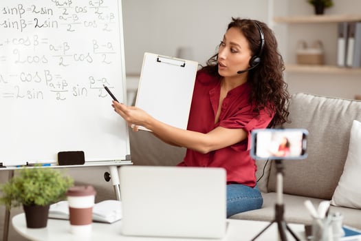 E-Learning. Confident woman having conference call using laptop. Lady teacher in headset showing clipboard, recording tutorial with mobile phone on tripod, streaming live pointing at board photo