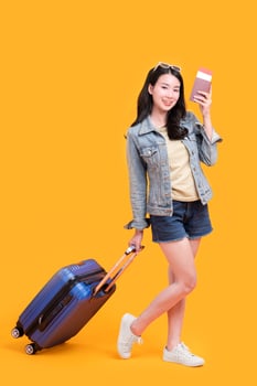 Happy young Asian tourist woman holding passport and boarding pass with baggage going to travel isolated on orange background. photo