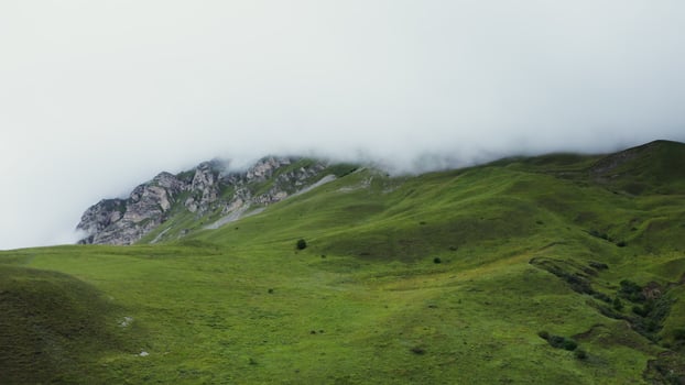 Hilly terrain, overgrown with green grass with rocky mountains, the tops of which are covered with thick clouds. View of the mountain valley from a quadcopter photo
