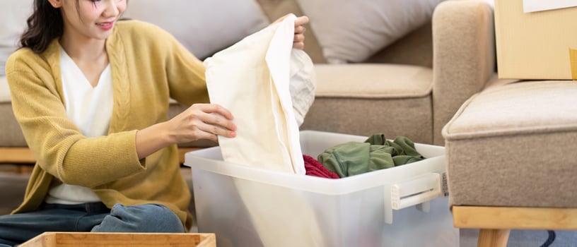 Smiling woman folding clothes for donation from a clear bin in her warm living room, promoting kindness through organization photo