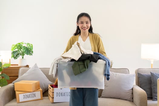 Smiling woman holding a transparent box full of clothes for donation, standing in a cozy living room photo