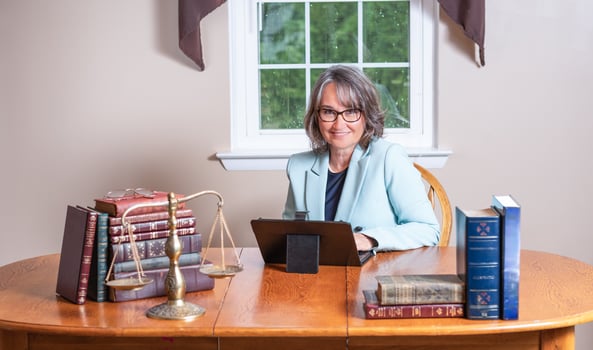 Female lawyer wearing glasses and a blue coat sitting at a laptop at a table between stacks of books and a balance scale with a window in the background. photo