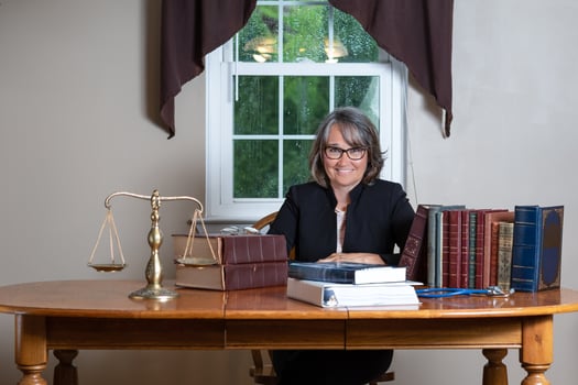 Woman attorney wearing glasses and a black coat sitting at a table behind stacks of legal books, a balance scale, and a stethoscope in front of a window smiling. photo