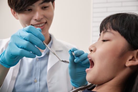 A young Asian male pediatric dentist checks and examines a little Thai girl's teeth in dental clinic, well-being mouth hygiene, and professional orthodontic healthcare work in a kid hospital. photo