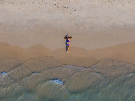 Aerial perspective of a tranquil beach scene with gentle waves meeting the sand. A person in a blue swimsuit lies near the shoreline, surrounded by calm. photo