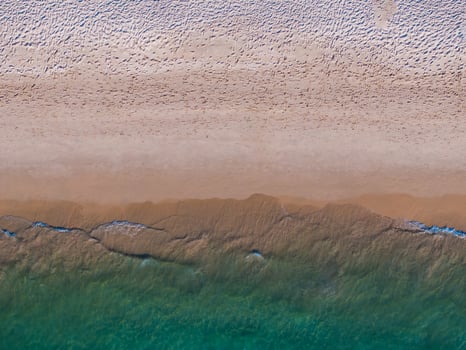 Aerial image of a sandy shoreline with visible footprints, wet sand, and greenish blue ocean waves gently meeting the beach in Thailand. photo