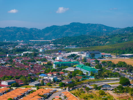 Aerial view of a town in Phuket, Thailand, featuring red tiled roofs, green hills, modern buildings, and a clear blue sky in the background. photo