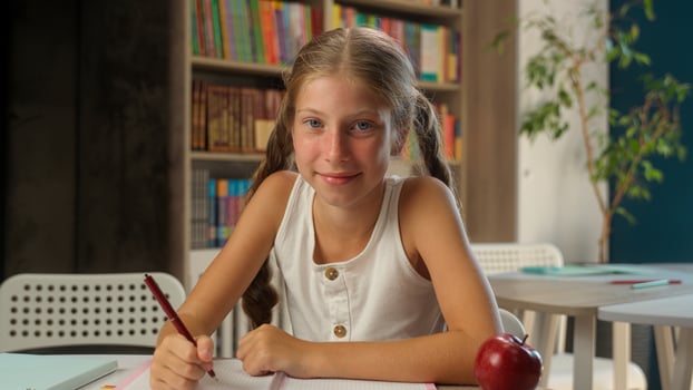 Little Caucasian girl writing copybook eating apple snack break lunch child smile schoolgirl study learning primary education school learner pupil female kid wink bite student healthy food fruit desk photo