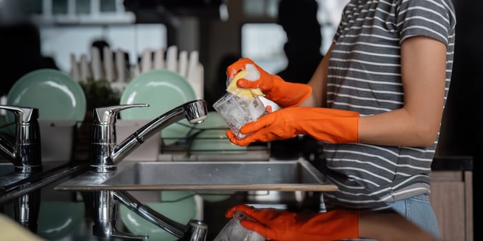 Young woman scrubbing a glass with sponge at kitchen sink, wearing orange gloves and smiling photo