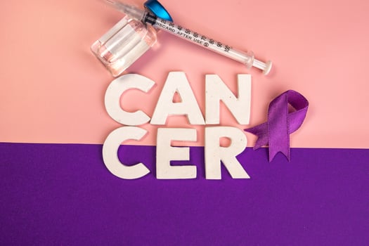 The word cancer is spelled out with white letters next to a purple ribbon and a syringe. World cancer day concept photo