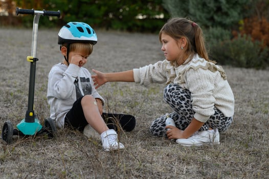 The boy, teary-eyed, sits next to his scooter while a caring girl sprays antiseptic on his injury, offering support. photo