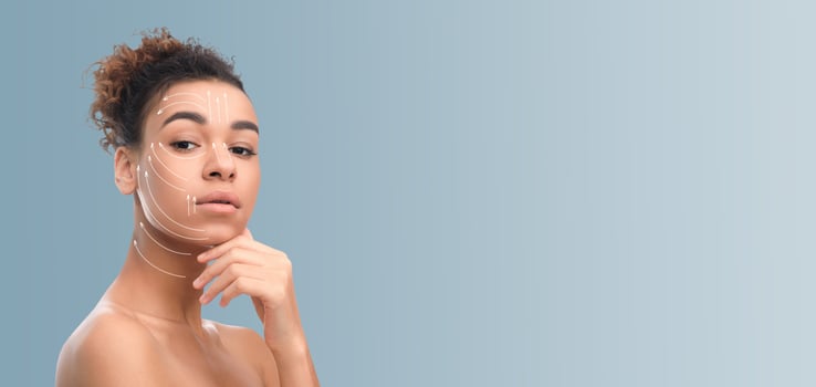 African American woman with curly hair is engaged in her skincare routine, gently touching her chin while displaying a calm demeanor. The background features neutral tones photo