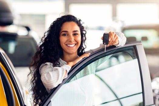 Cheerful brunette woman buying new cute car, raising hand up with automatic key and smiling, showroom interior, copy space. Pretty curly lady customer purchasing yellow female auto, getting in photo