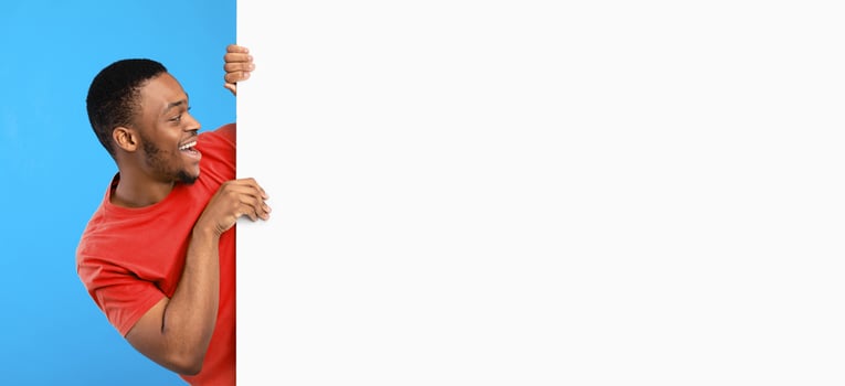 Discount Offer. Excited Black Man Looking At Empty White Board Posing Near Big Poster With Free Space Over Blue Background. Advertisement Banner With Man Advertising Your Text. Mockup, Panorama photo