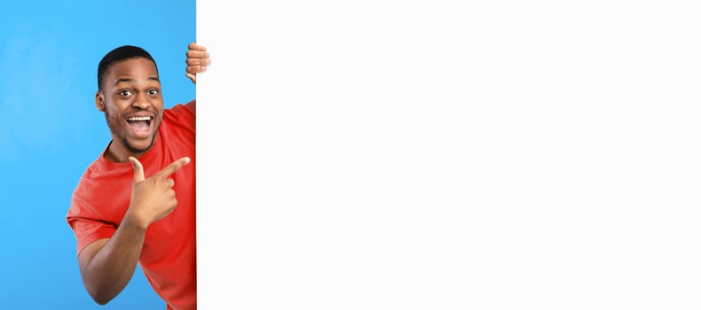 Great Sales Offer. Excited African American Man Pointing Finger At Copy Space On Big White Paper Poster Advertising Your Text Over Blue Studio Background, Smiling To Camera. Panorama, Mockup photo