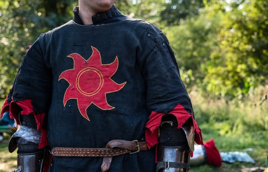 a man in the costume of a medieval knight. his face is not visible. a red sun is embroidered on his medieval shirt. the photo was taken vertically and large. medieval shirt of black and red color