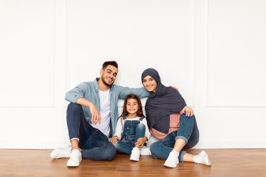 Portrait of cheerful arabian man, woman in headscarf and cute girl sitting on the floor leaning on white wall posing in new apartment in empty living room, family looking at camera, free copy space photo
