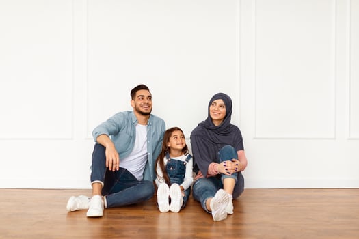 Portrait of smiling muslim people sitting on floor leaning on white wall in empty apartment, family thinking about new house design after moving, looking up and aside at free copy space photo