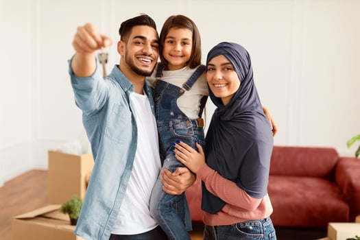 Portrait Of Happy Muslim Family Showing Keys, Cheerful Parents Hugging Little Kid, Holding Daughter On Hands. Arabic People Moving In New Apartment Standing In Living Room. Selective Focus photo