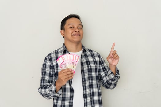 Asian guy smiling happy while holding mobile phone and bunch of money in Rupiah currency. photo