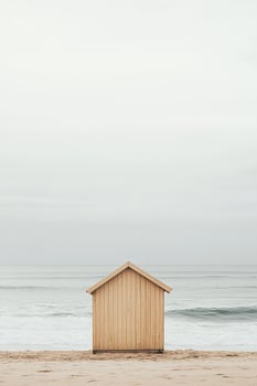 A minimalist wooden beach hut in neutral tones stands alone on a smooth sandy beach. Soft waves gently lap the shore, creating a serene and modern atmosphere perfect for relaxation photo