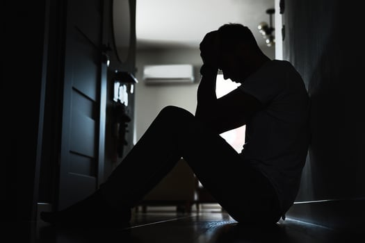 Depressed young man sitting on floor in dark room. Silhouette of young male suffering from loneliness and anxiety, covering his face with hands. Person with mental disorder photo