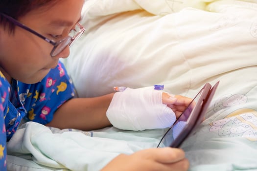 Little Asian boy illness lay on sickbed in hospital with a saline intravenous in hand holding smartphone and watching on youtube. photo