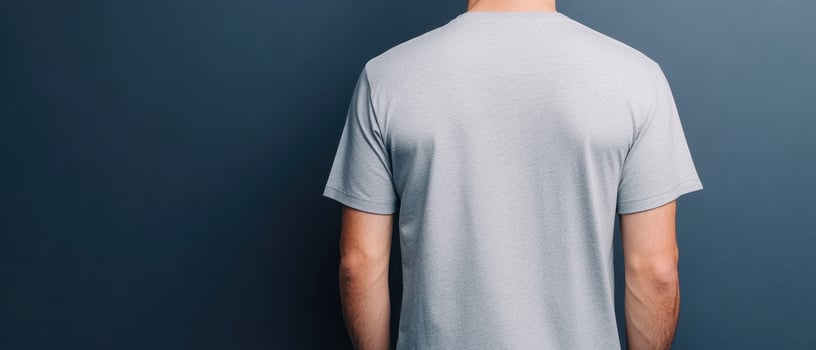 man wearing grey t shirt stands against dark blue wall, showcasing back view of shirt. simple design highlights fabric texture and fit, creating casual and modern look photo
