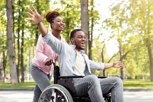 Happy black disabled guy in wheelchair on walk with his loving girlfriend outdoors, having fun, spending time together. African American impaired couple enjoying each other's company at green park photo