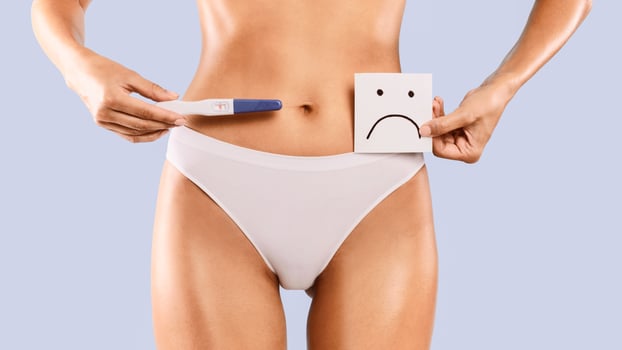 Unplanned Pregnancy. Young woman holding positive pregnancy test with two lines and sticker post-it note card with unhappy sad smiley face. Unrecognizable female in white panties. Closeup cropped view photo