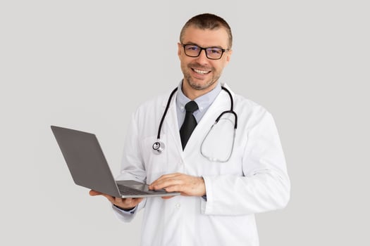 Online Consultation Service Concept. Portrait Of Smiling Confident Doctor In Glasses Using Laptop, Standing Isolated On White Studio Background, Copy Space. Physician wearing coat and stethoscope photo
