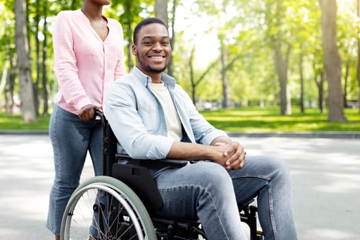 Happy black woman taking her impaired boyfriend in wheelchair for walk at city park. African American disabled couple enjoying nature together, spending time outdoors photo