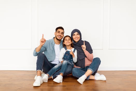 Portrait of smiling muslim people sitting on the floor leaning on white wall in new apartment, looking pointing fingers up. Cheerful man, woman and girl planning renovation design, showing copy space photo