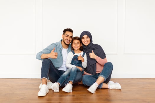 Happy Loving Family. Portrait of smiling muslim people sitting on the floor near white wall in new apartment. Cheerful man, woman in hijab and little girl showing thumb up sign gesture photo