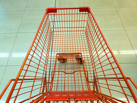 An empty orange shopping cart with a wireframe design is seen from above on a tiled floor. The cart is positioned in a well-lit, clean environment, likely a supermarket or store photo