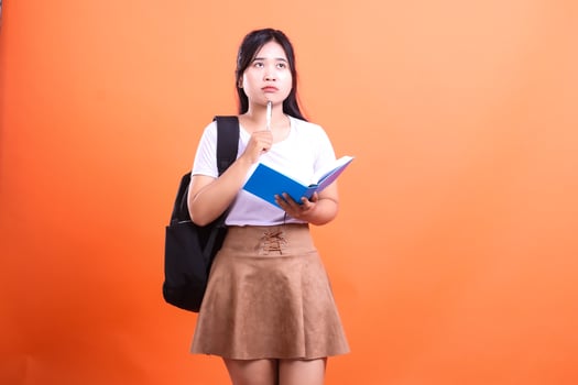 A young student in a white t-shirt and brown skirt stands against an orange background while holding a pen and book photo