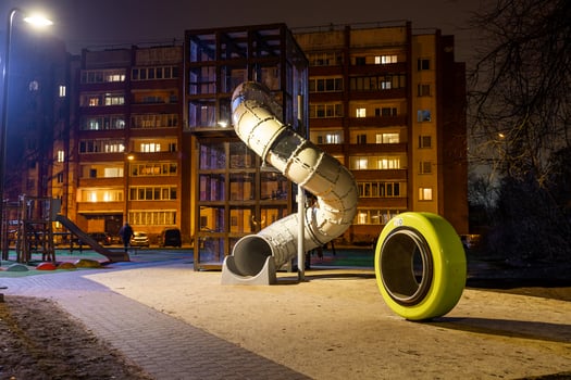 Nighttime view of a playground with a tall spiral slide, yellow play element, paved walkway, and a lit residential building in the background. photo