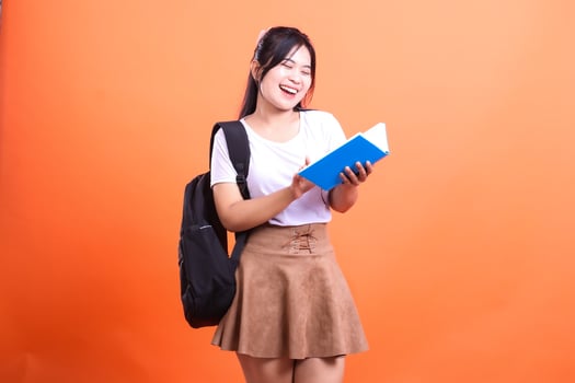 Smiling student in white shirt and brown skirt holding a blue book with a black backpack on an orange background photo
