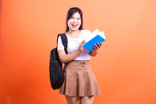 A student with a white t-shirt, a skirt, and a black backpack happily holds a blue book in front of an orange background photo