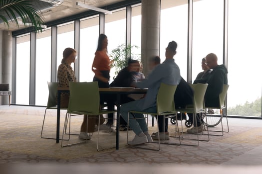 A diverse business team, working urgently around a conference table, collaborates on a meeting while their director, seated in a wheelchair, waits for their input photo