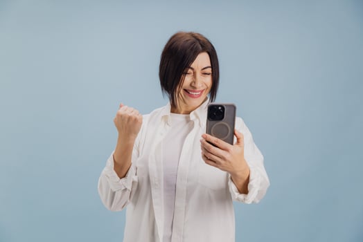 Excited mature woman celebrating success while using smartphone on blue background photo