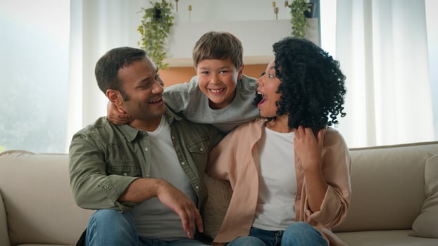 Affectionate parents adult African American mother and father embrace cuddle on couch funny little boy adopted son kid child jumping behind hug mom and dad laughing multiracial family portrait at home photo