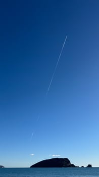 A clear blue sky with a long contrail stretching above a small island on calm sea waters . copy space photo