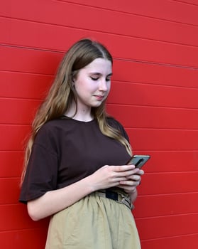 young woman with long, light brown hair is standing in front of a bright red wall, looking down at her phone. photo
